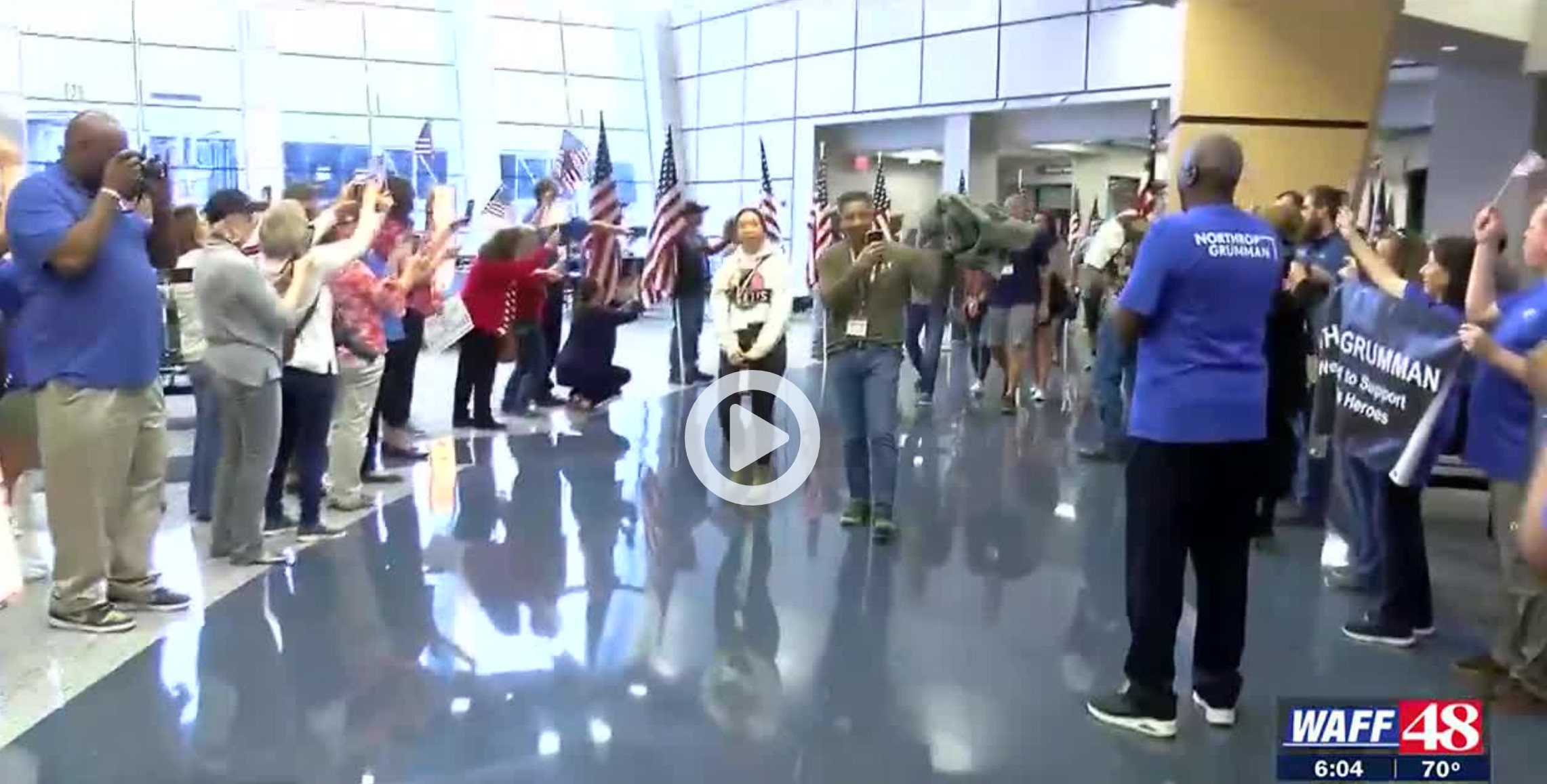 Screenshot of paused video coverage from WAFF 48 News of Veterans and caregivers arriving at the Huntsville International Airport to participate in Heroes Week 2022; shows people gathered and cheering along the walkway near the TSA exit, some holding large flags in a flagline, others taking photos of the incoming participants; and some incoming heroes are seen walking toward the camera. WAFF 48 logo and time stamp 6:04 PM in the bottom right corner.