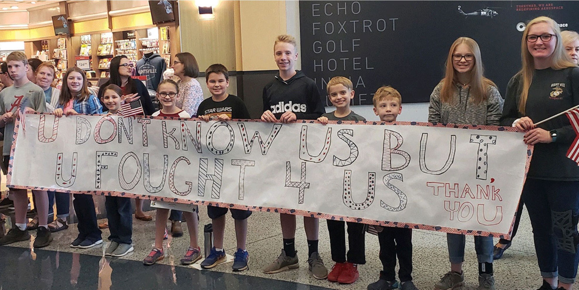 Semper Fi Community Task Force photo taken inside the Huntsville International Airport, shows children holding a long banner that says "You Don't Know Us But You Fought 4 Us" - to help welcome the Veterans and Caregivers participating in Heroes' Week 2022